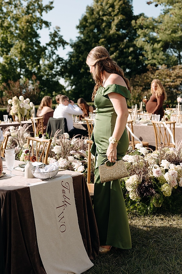 Wedding guest attire in a green satin bridesmaid dress with a gold clutch and wrist corsage beside garden reception tables on a lawn