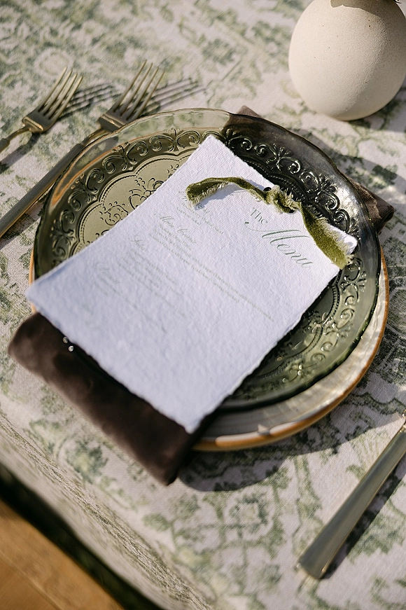Wedding place setting with a wedding menu card on a folded napkin tied with olive green ribbon, gold flatware and embossed charger plate on wood tabletop