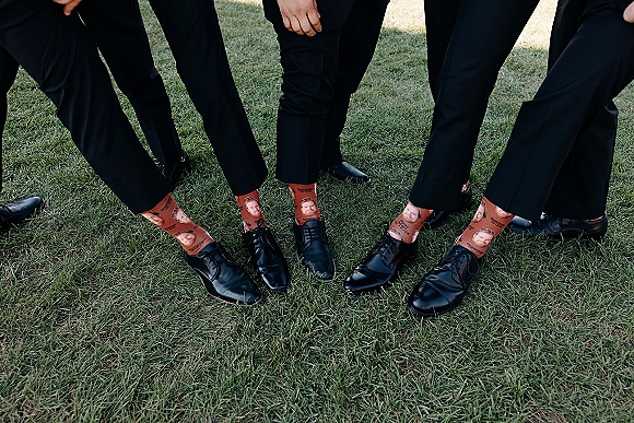Groomsmen socks and black dress shoes on a grass lawn, showing printed funny groomsmen socks beneath black suit pants