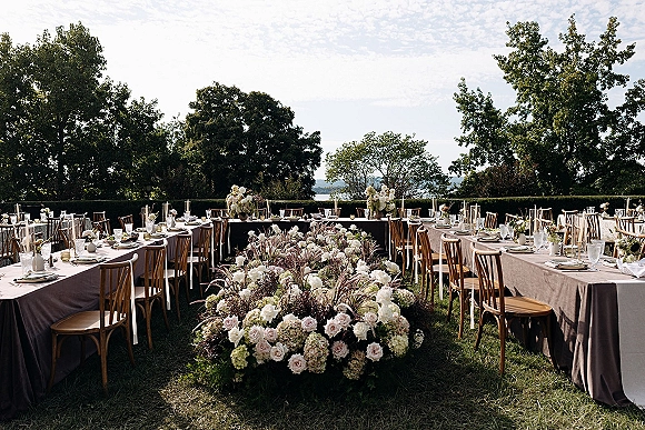 Outdoor reception tablescape with round and rectangular tables in taupe linens, wooden chairs, tall taper candles, and rose-hydrangea florals on a lawn near water view