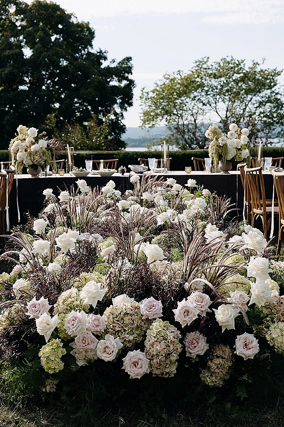 Reception tablescape with outdoor reception tables on long black tablecloth banquet setup, roses, hydrangeas, taper candles on a lawn by water