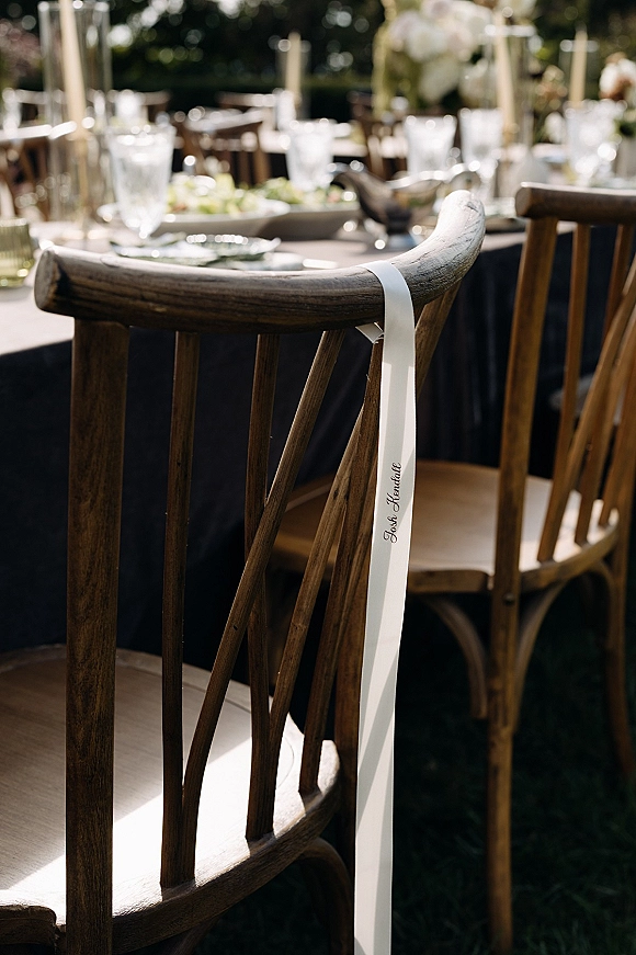 Wedding place cards tied with a chair place card ribbon on wood chairs, framing an outdoor reception tablescape with candles and florals