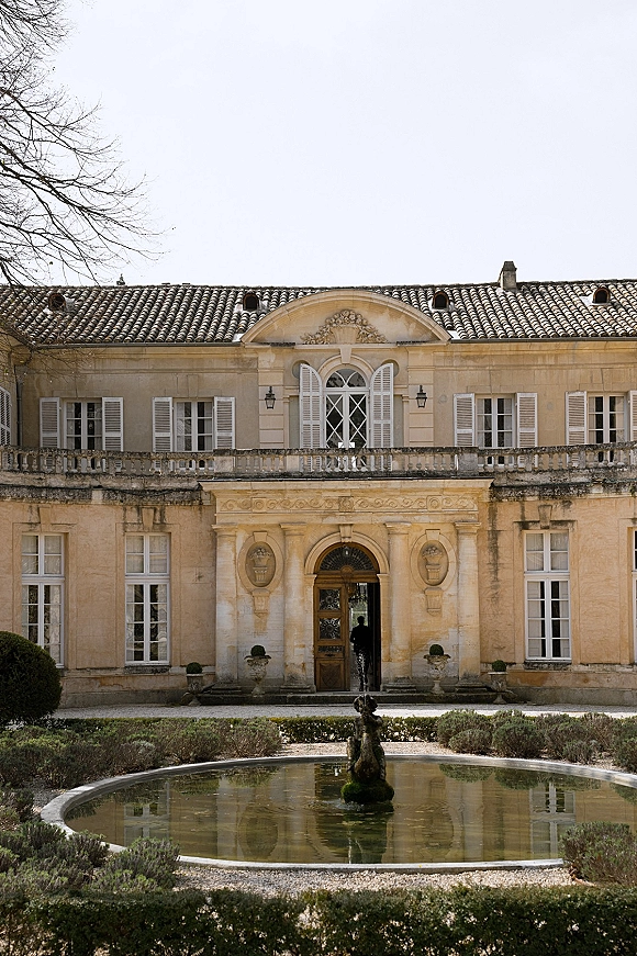Wedding venue exterior with a stone fountain and shuttered estate facade, set in a manicured garden and gravel courtyard under open sky