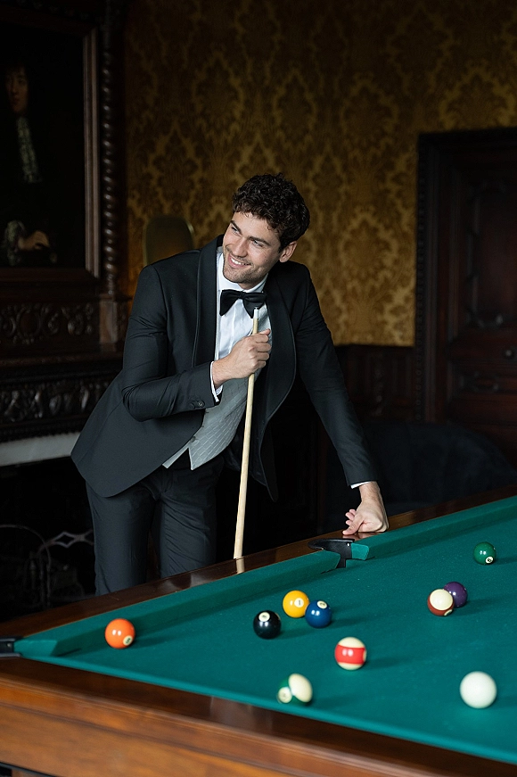 Groom portrait in black tie tuxedo holding a pool cue beside a pool table, set in a vintage lounge with leather sofa and wallpaper.