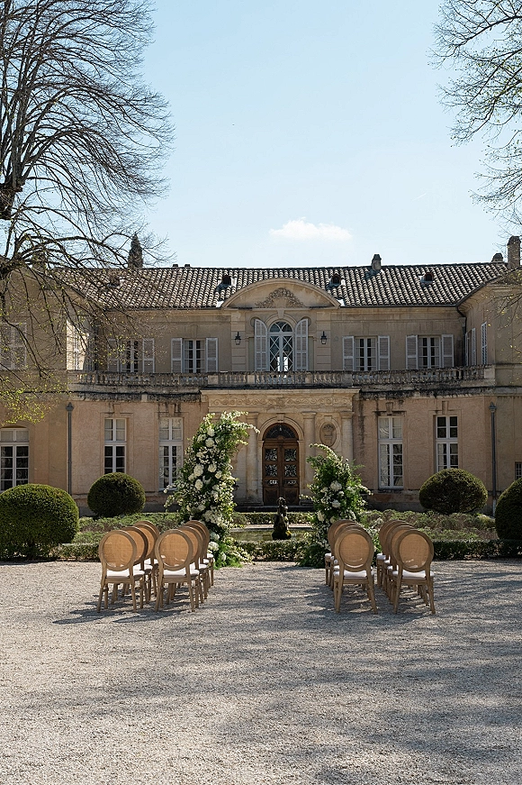 Ceremony setup for an outdoor wedding ceremony with natural wood chairs lining a gravel aisle toward white floral arches in an estate courtyard