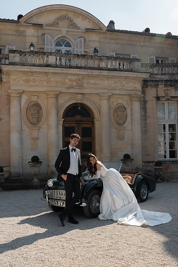 Couple portrait of bride and groom with car, her long satin dress train by a vintage convertible in a mansion gravel courtyard.