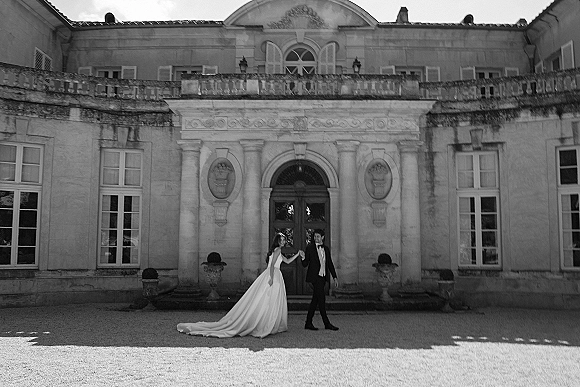 Couple portrait of bride and groom holding hands, her long train gown and his black tuxedo framed by a stone villa entrance.
