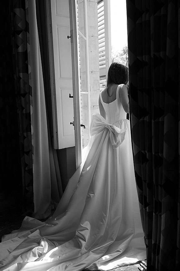Bridal portrait of a bride looking out window, showing a backless satin wedding dress with an oversized bow and long train in soft window light