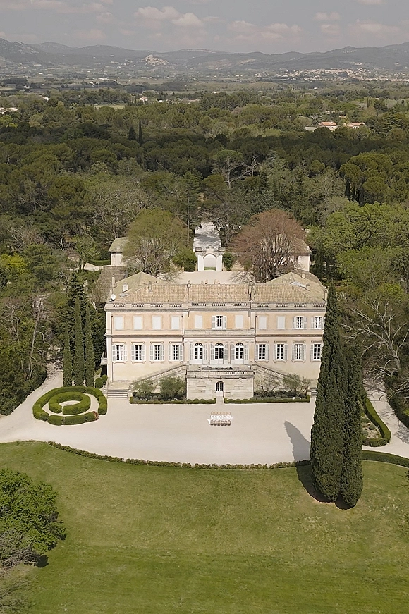 Wedding venue exterior featuring a stone mansion and gravel courtyard with manicured hedges, framed by forest and distant mountains