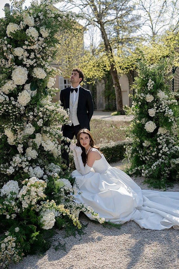 Couple portrait of bride in opera gloves and cathedral train seated by groom in tuxedo beneath a white hydrangea arch in a garden path