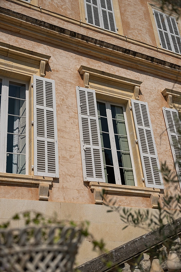 Wedding venue exterior with tall windows and white shutters on a sunlit stone facade, framed by tree branches and ledges