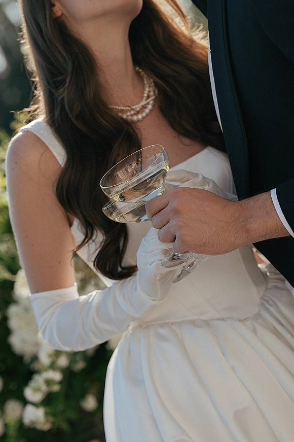 Wedding champagne toast as bride in satin dress and gloves with pearl necklace clinks coupe with groom in tux amid greenery and white flowers