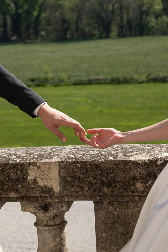Couple holding hands in a wedding handholding photo, groom in suit jacket and bride in white dress by a stone balustrade and lawn