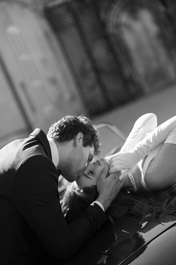 Wedding kiss in a black and white wedding photo as groom dips gloved bride on a car hood, pearl bracelet visible against building facade
