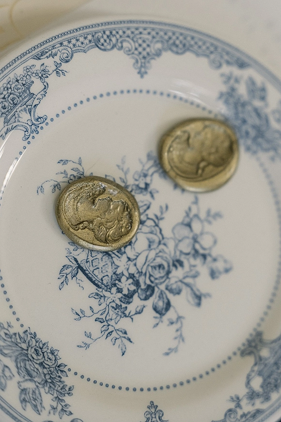 Wax seals in gold wax seal designs arranged on a blue and white floral china plate, captured as a wedding stationery detail flat lay