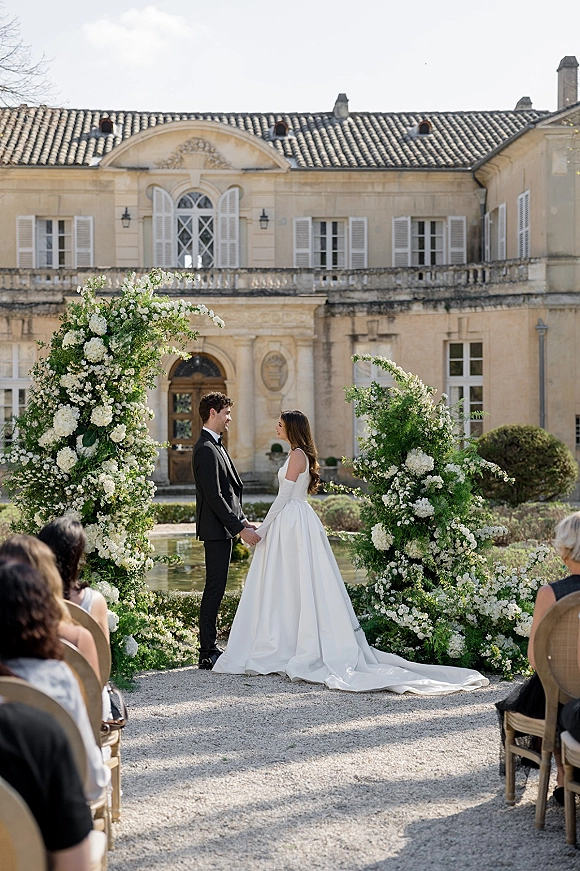 Wedding ceremony moment outdoors as bride and groom hold hands under a white floral arch along a gravel aisle in an estate courtyard garden