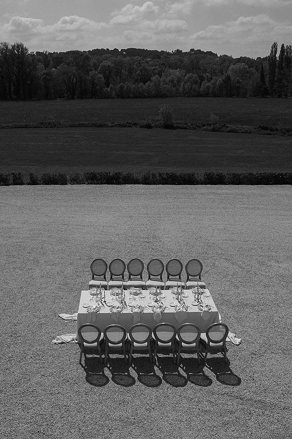 Outdoor reception table set with a long banquet table, white tablecloth, place settings, and wine glasses on a meadow lawn under cloudy sky