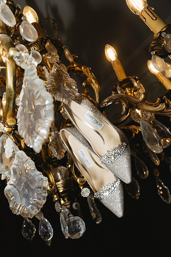 Wedding shoes perched on a crystal chandelier, sparkly heels with rhinestone embellishment against a dark ceiling and gold frame