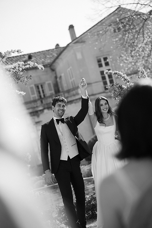 Recessional moment as bride and groom smile with raised hands, strapless gown and bow tie, in garden by an estate building