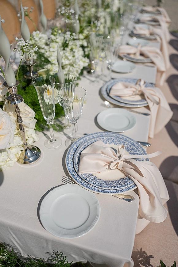 Reception tablescape with wedding place setting featuring blue patterned plates, blush ribbon-tied napkins, white taper candles on silver candlesticks outdoors