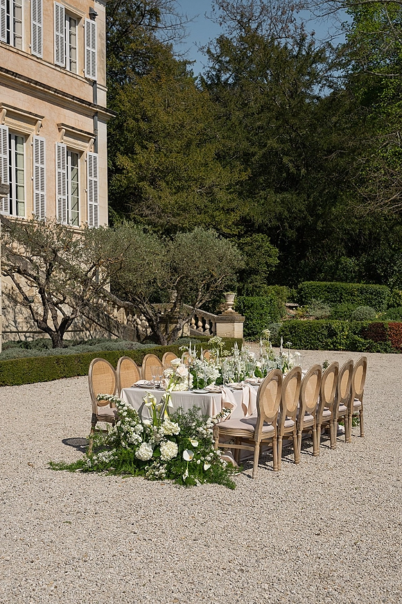 Outdoor reception table set for an al fresco wedding dinner with white linens, blush runner, taper candles, and white florals in a stone estate courtyard