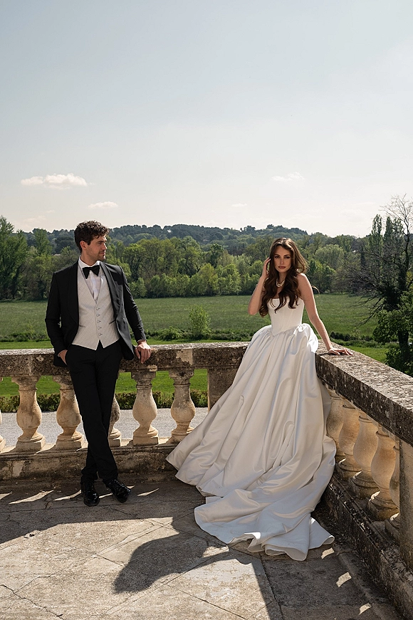Couple portrait of bride in a strapless ball gown with pearl necklace and groom in black tuxedo on a stone terrace overlooking countryside fields