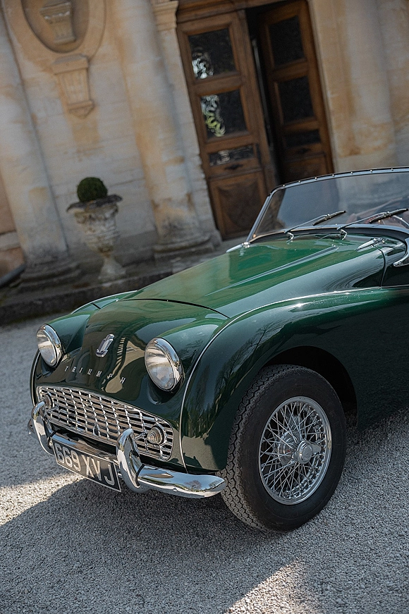 Wedding getaway car, a green vintage convertible with chrome bumper and wire wheels parked by stone columns and wooden doors