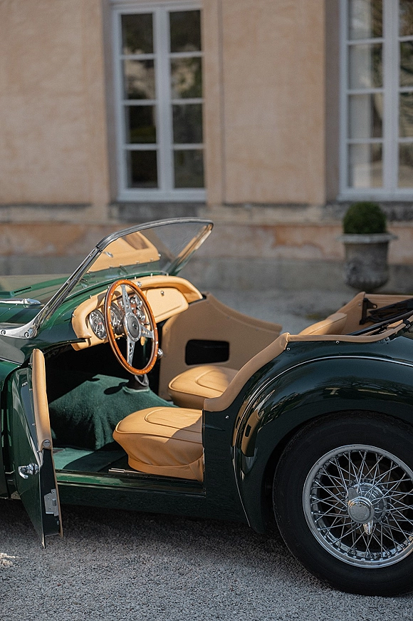 Vintage wedding car parked with door open, showing tan leather seats and wood steering wheel on a gravel drive by a stone facade