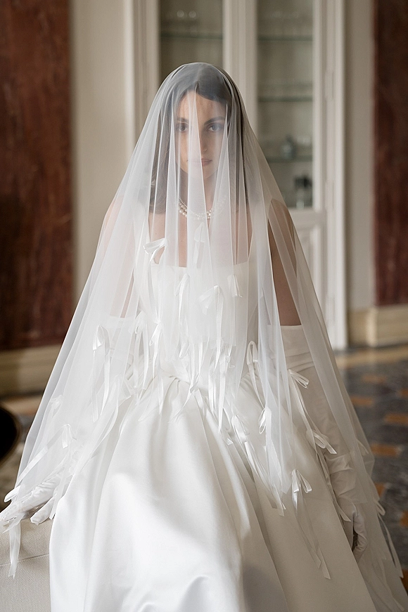 Bridal portrait of a bride with veil over face, wearing a strapless satin gown and long gloves, seated in a wood-paneled room.