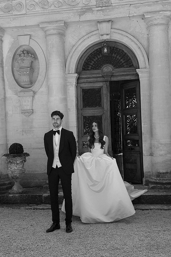 Couple portrait in formal wedding attire on stone steps, bride in strapless ball gown beside groom in tuxedo at arched doorway