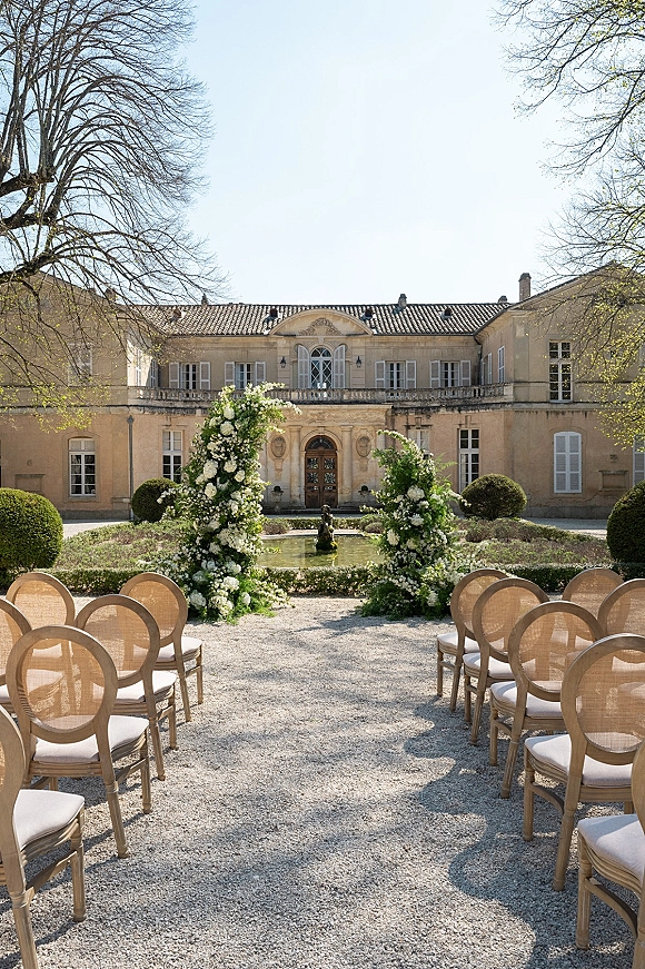 Ceremony setup with aisle floral pillars of white flowers and greenery lining a gravel aisle toward a fountain in a mansion courtyard garden