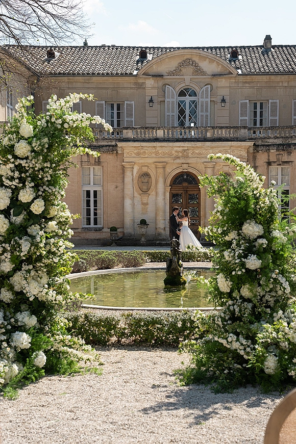 Couple portrait of bride and groom kissing under a white floral arch, holding hands in a courtyard garden before a historic mansion