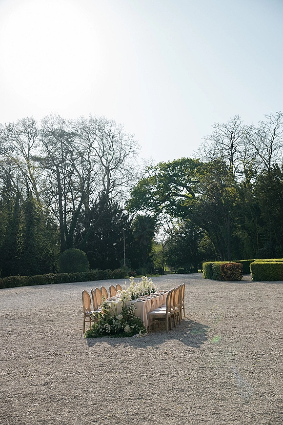 Outdoor reception table with a long banquet table wedding setup, white linens and floral garland centerpiece in a sunlit gravel courtyard