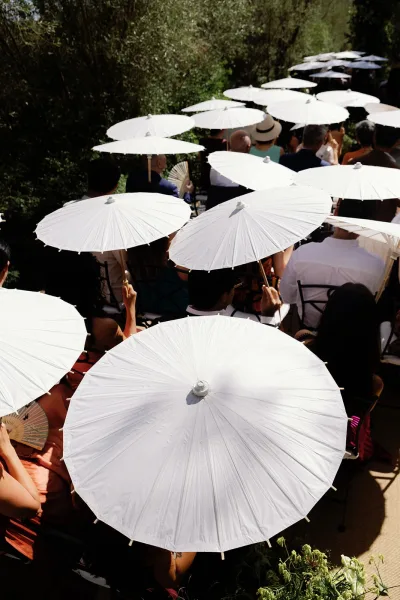 Ceremony parasols line the aisle as wedding guest parasols, with hand fans provided beneath garden trees.