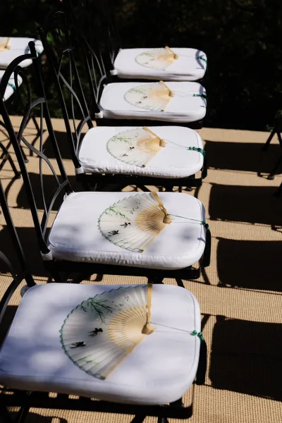 Ceremony seating with outdoor wedding chairs arranged in rows, each set with a paper hand fan, on woven mat flooring amid lush garden greenery.