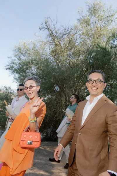 Wedding guests in outdoor wedding guest attire walk a tree-lined dirt path under a blue sky, some wearing sunglasses in the summer sun.
