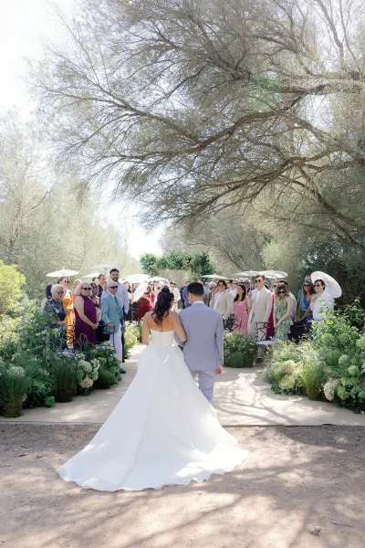Ceremony processional with the bride walking down the aisle arm in arm with the groom as guests shade with parasols along a tree-lined garden path.