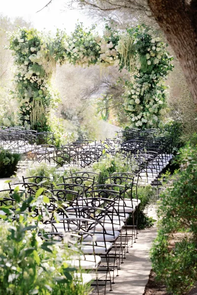 Ceremony setup featuring outdoor ceremony decor with a floral arch framed by garden trees and shrubs beside a garden walkway.