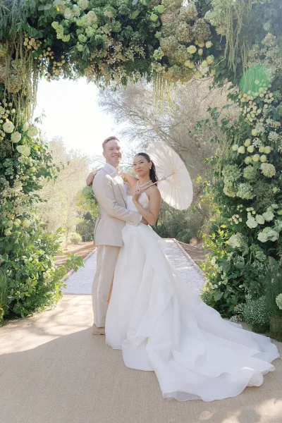 Couple portrait of a bride and groom under a green and white floral arch in a garden, standing on a gravel path amid garden trees.