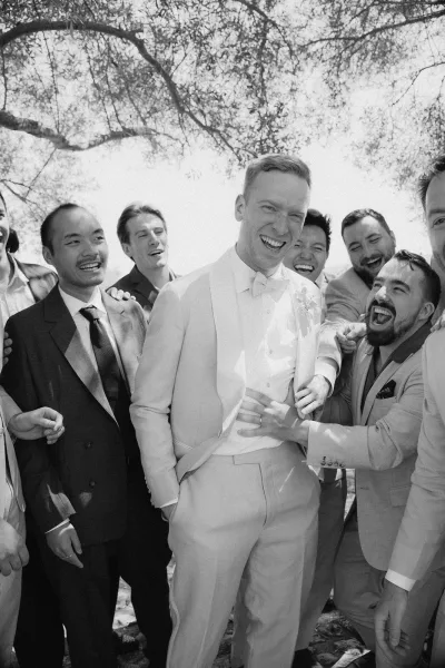 Groom and groomsmen share a relaxed wedding party portrait beneath a tree canopy, highlighted by the groom’s classic bow tie.