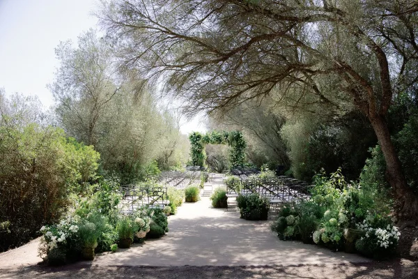 Outdoor ceremony setup along a garden ceremony aisle, centered on a rectangular floral arch in lush green and white blooms.