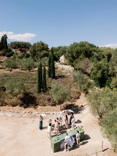 Welcome drink station for an outdoor cocktail hour, with paper parasols for guests beneath olive and cypress trees along a sunlit hillside.