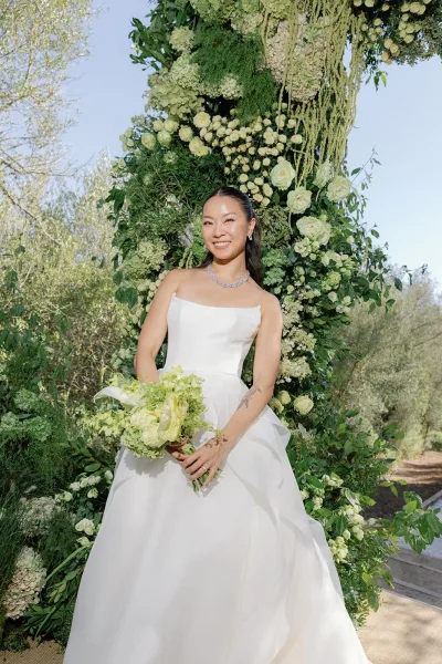 Bridal portrait of a bride holding a bouquet along a garden pathway framed by trees and blue sky, capturing her serene wedding day look.