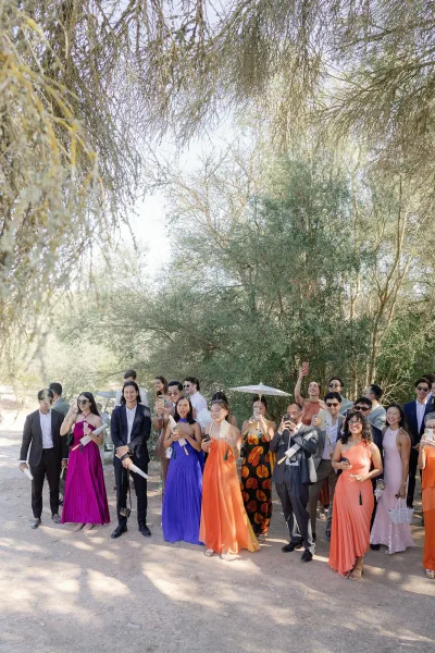 Wedding guests, guests cheering with hand fans, line a sunlit dirt path beneath trees, their excitement bright against the warm afternoon.