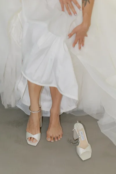 Bridal shoes: ivory block-heel sandals in an open-toe wedding style, accented by a crystal anklet on a concrete floor.