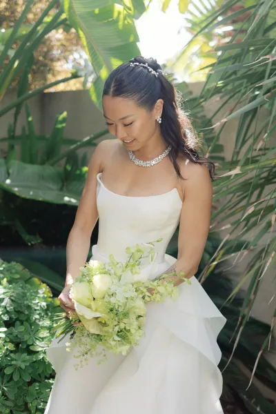 Bridal portrait of a bride holding bouquet, with a sleek ponytail, framed by palm leaves and a weathered stone wall in a tropical garden.