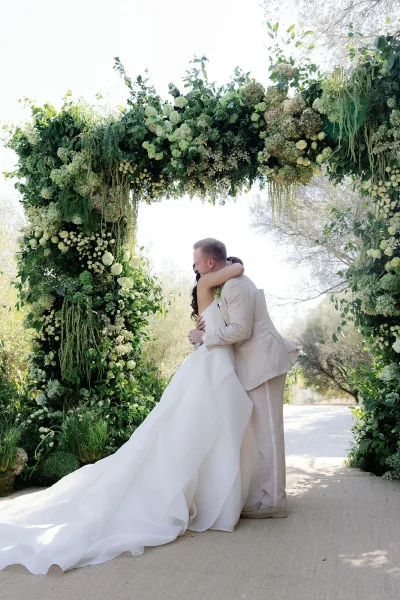 Ceremony moment as the bride and groom hug beneath a lush white-and-green floral arch, with garden trees lining the pathway.