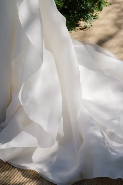 Wedding dress train with ruffled layers from a layered wedding gown, flowing across a woven rug with soft greenery framing the scene.