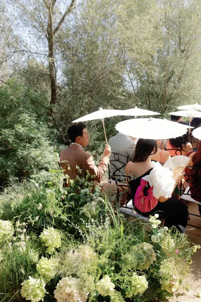 Ceremony parasols shade guests as thoughtful guest comfort ideas, with hand fans provided beneath garden trees and shrubs.