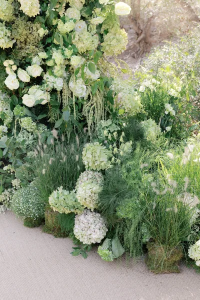 Ceremony floral installation of green and white ceremony flowers, highlighted by cascading amaranthus, set in a sunlit garden among shrubs and trees.